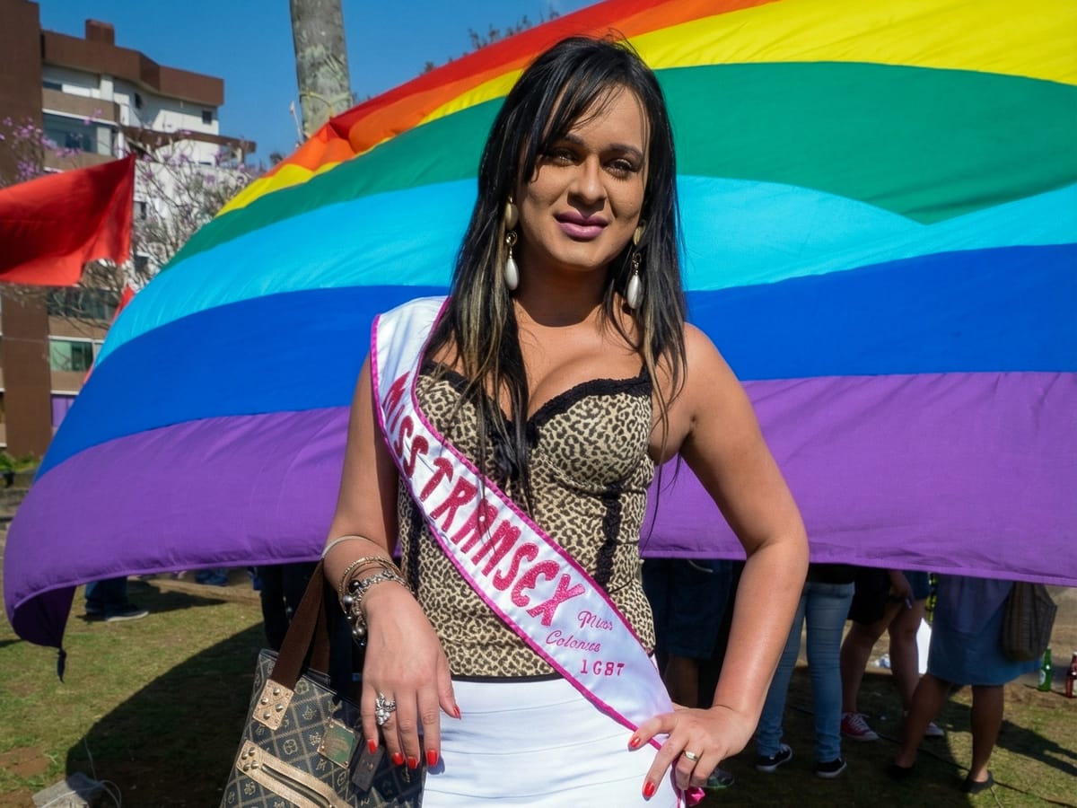 Participante com faixa de Miss Transex diante da bandeira LGBT em ação do Plural